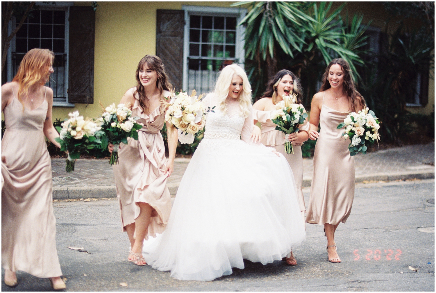 A bride and her wedding party cross a street in front of a yellow house.