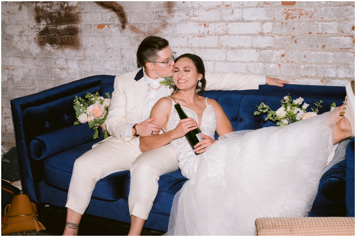A couple cuddles on a blue couch and holds a bottle of champagne in direct flash for wedding photography.