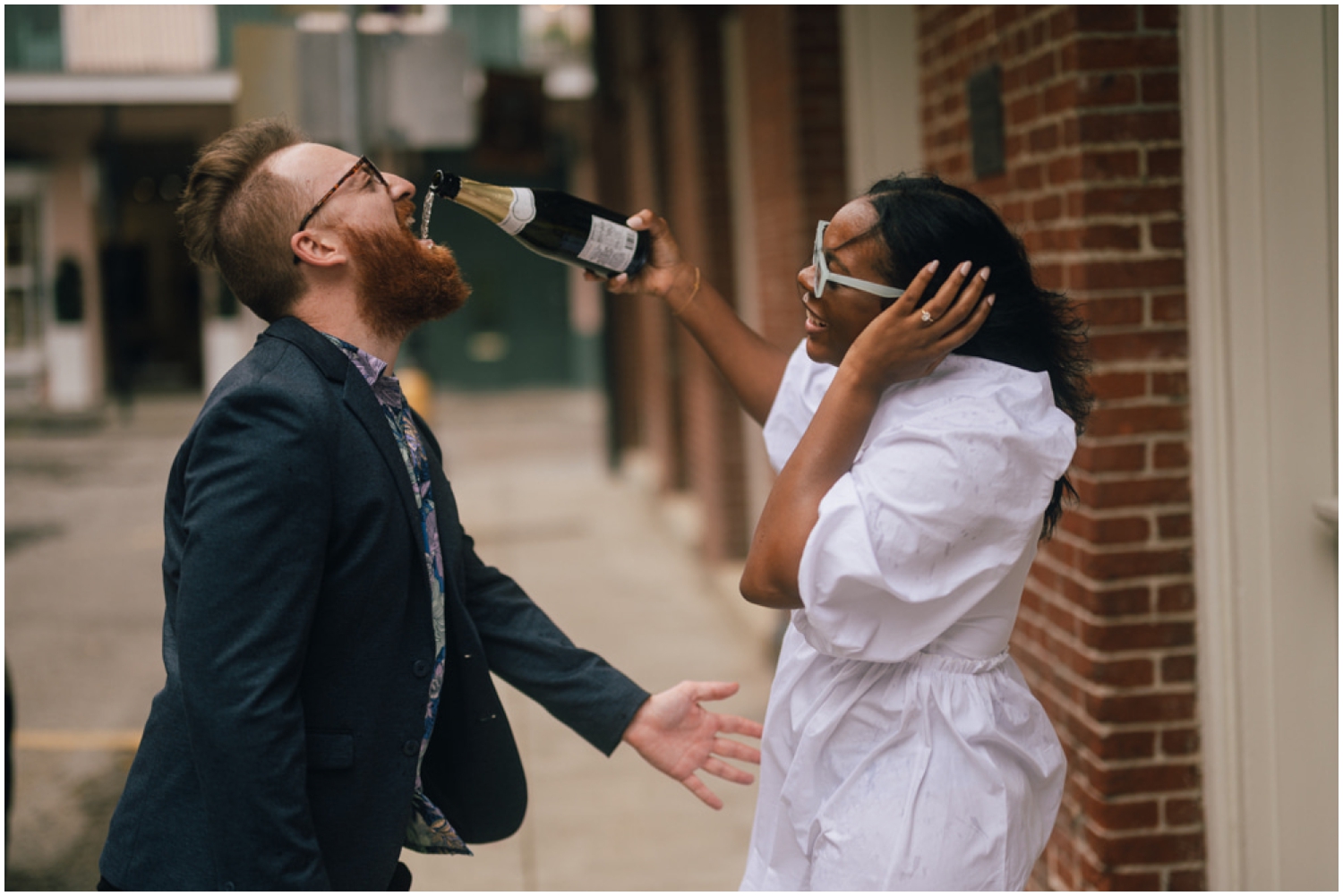 Paris pours champagne into Dan's mouth in front of a brick building in the French Quarter.