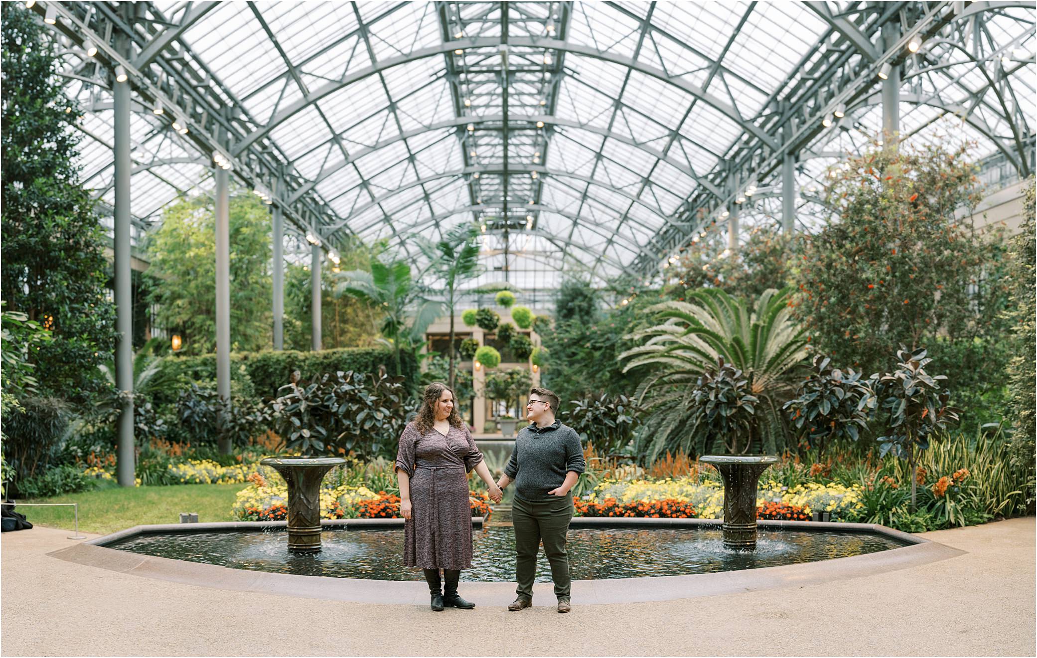 Two people hold hands in front of a conservatory fountain in Longwood Gardens engagement photos.