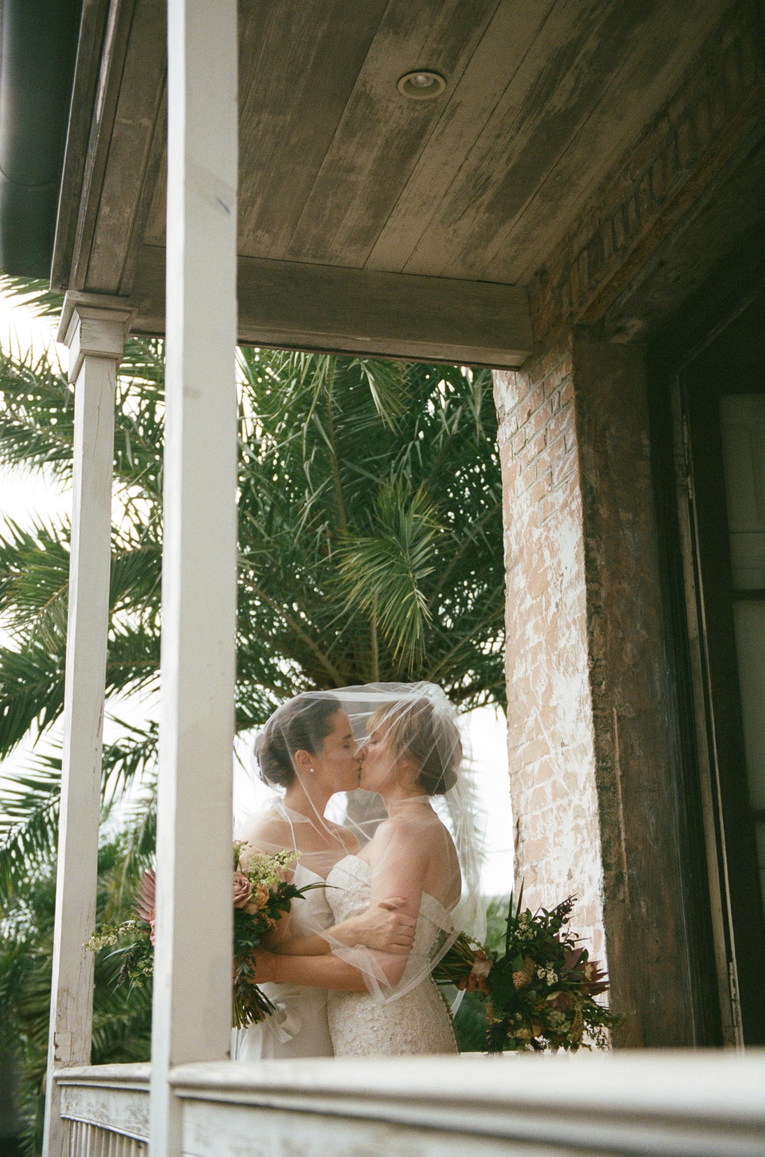 Two brides stand under a veil on a balcony and kiss above a Philadelphia wedding photographer.