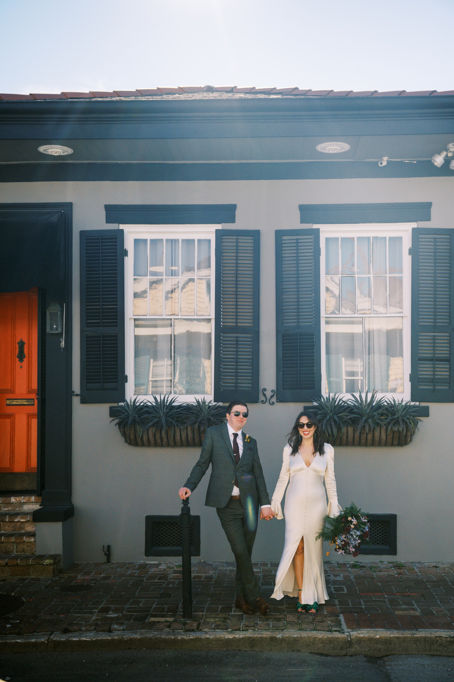 A bride and groom pose in front of a historic wedding venue wearing sunglasses.