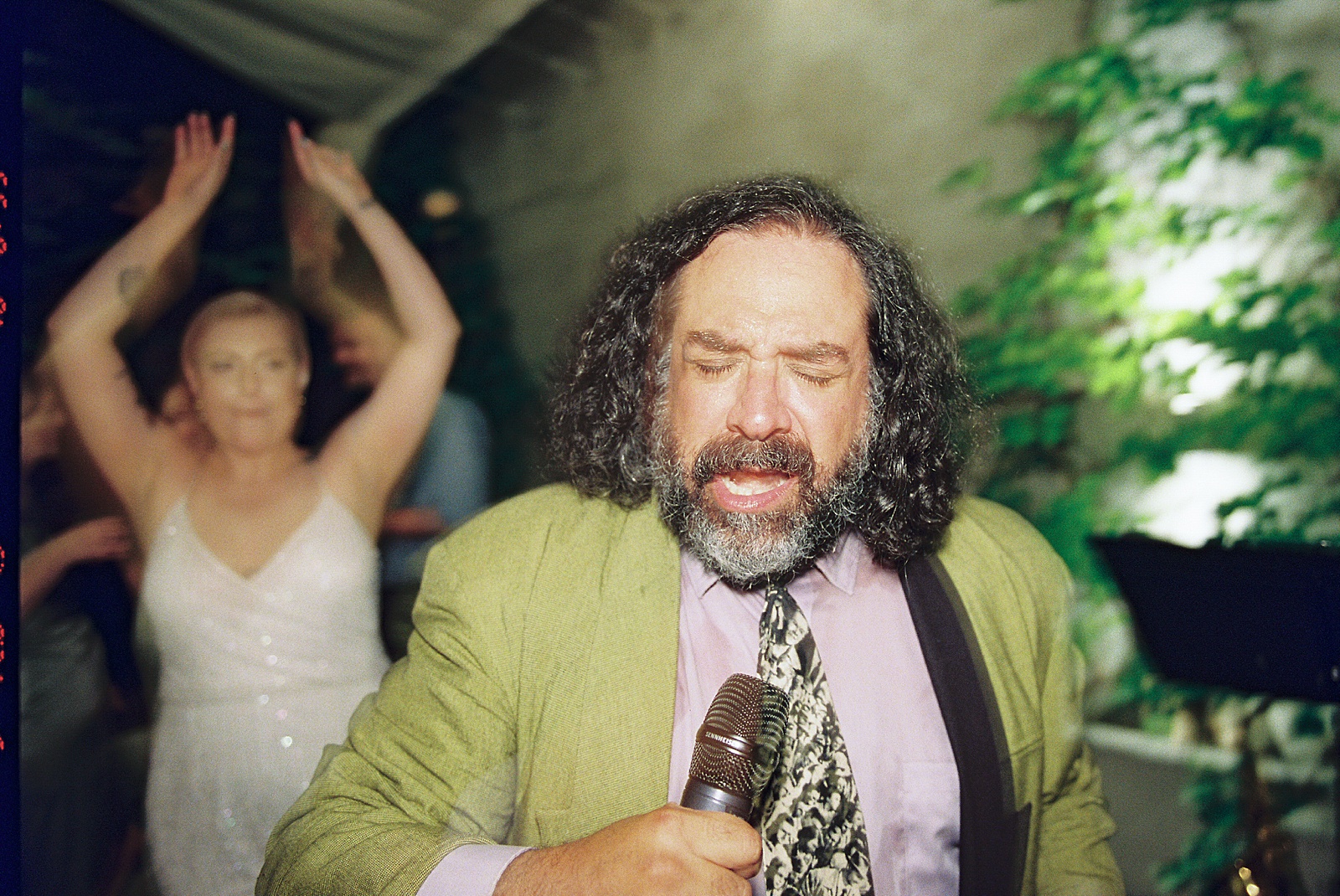 A wedding singer sings into a microphone while a bride dances behind him.