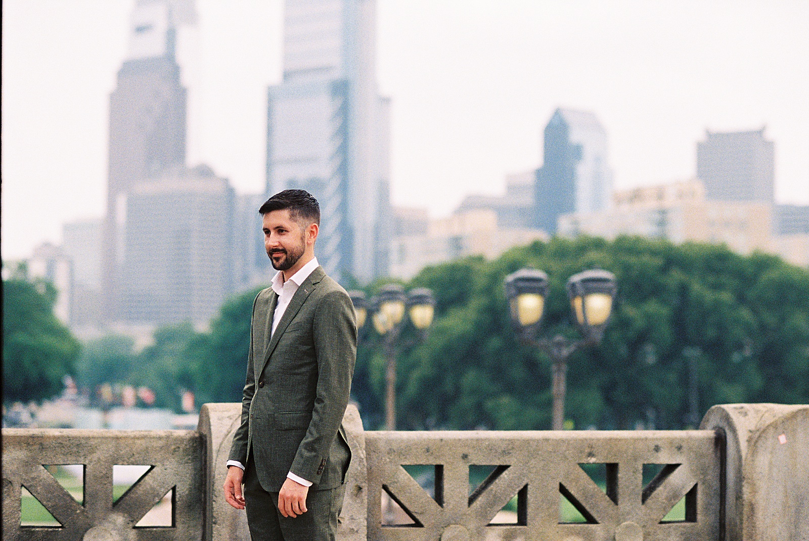 A groom stands on a Fairmount Park walkway with the Philadelphia skyline in the background.