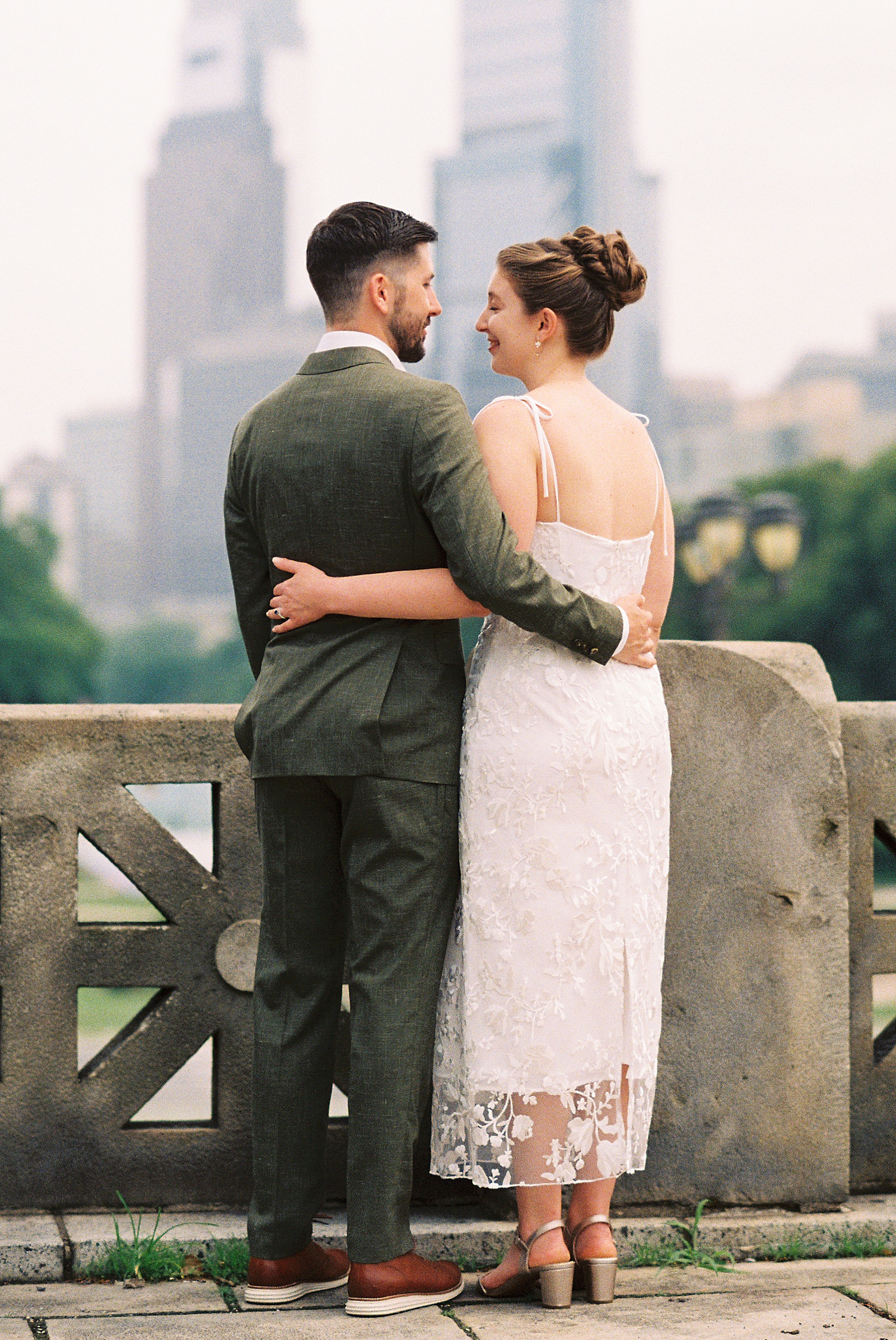 A bride and groom stand with their arms around each other's waists as they look at Philadelphia skyscrapers in the distance.