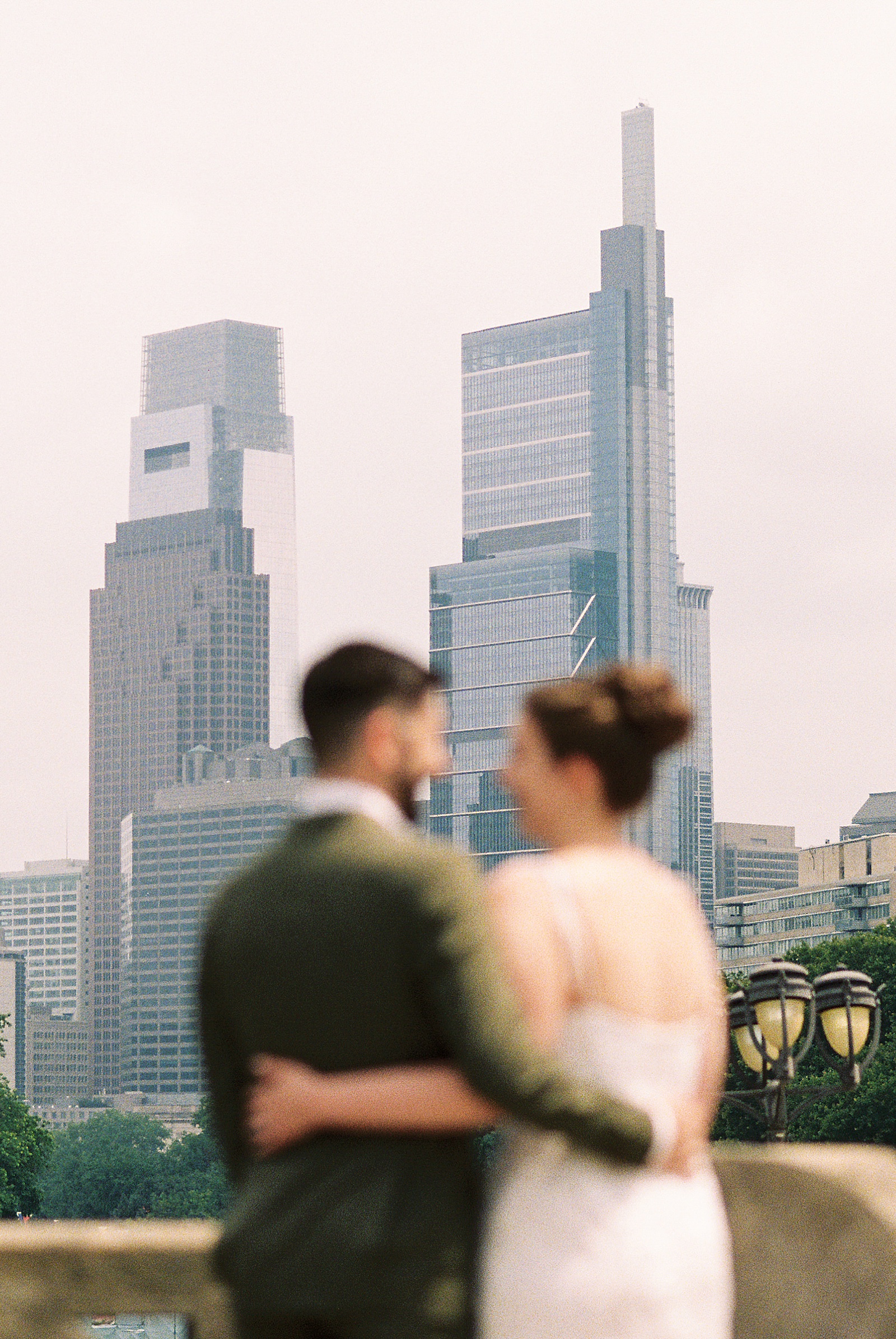 A film wedding photo shows a bride and groom out of focus in the foreground in front of the Philadelphia skyline.