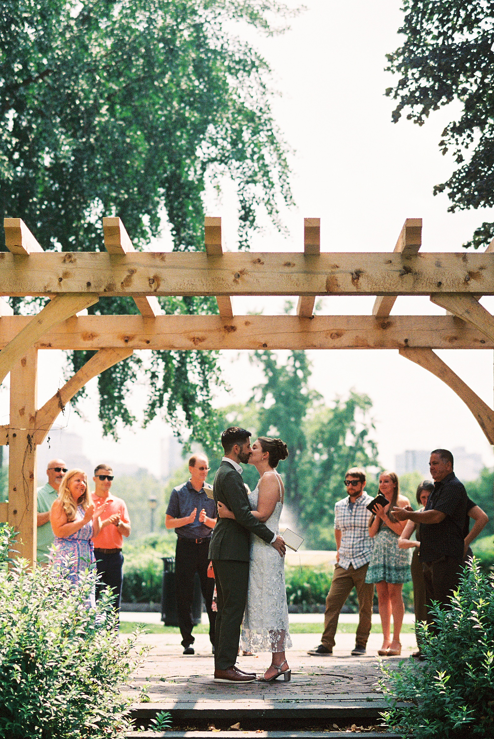 A bride and groom kiss at the end of their self-uniting ceremony while their families clap.