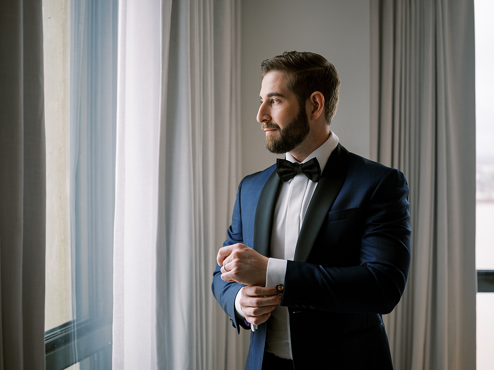 A groom buttons his cufflink while he looks out a window.