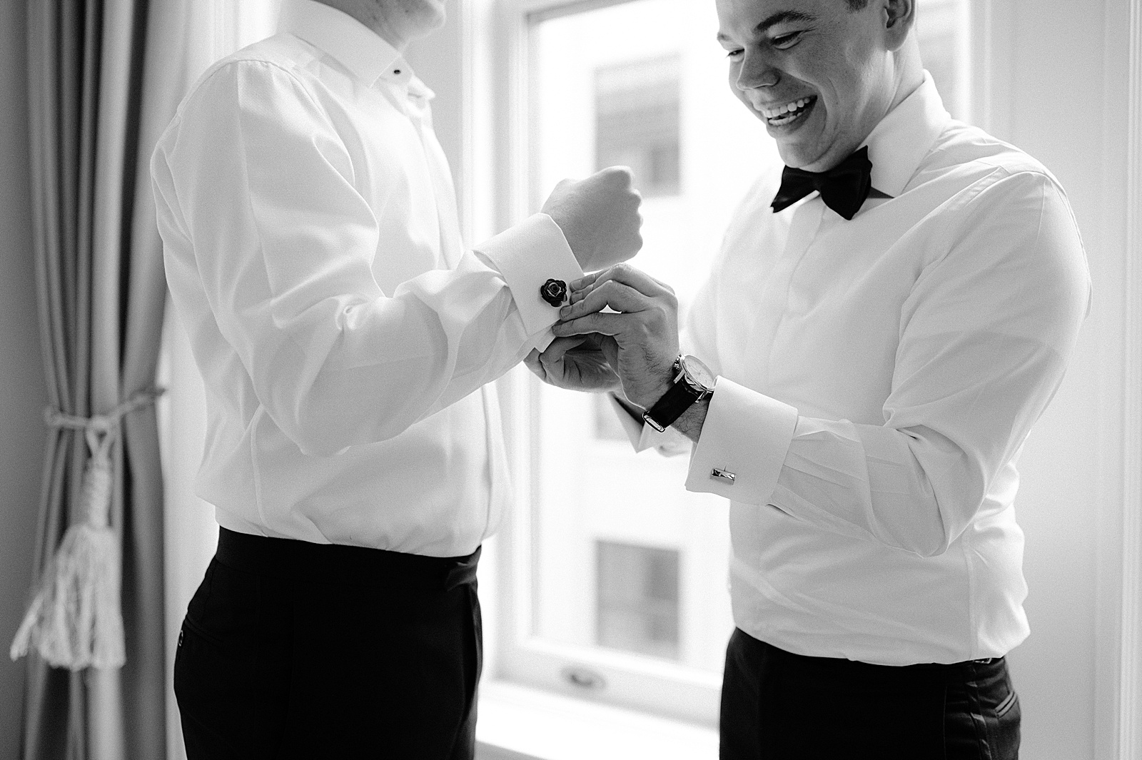A smiling groomsman helps a groom with his cufflinks.