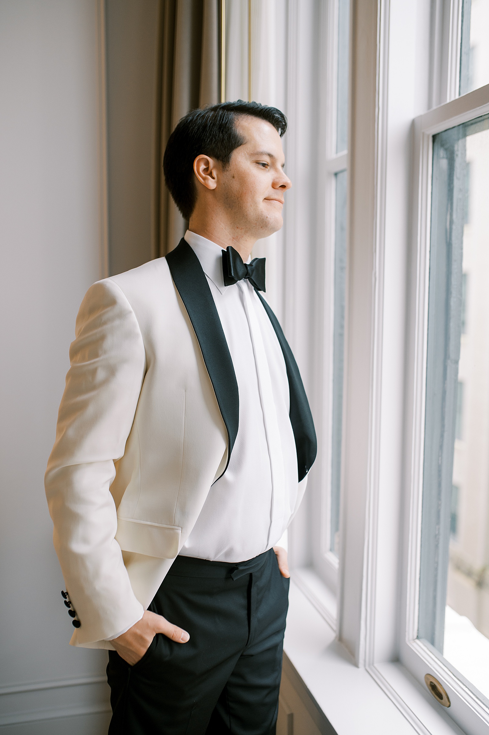 A groom in a white tuxedo poses for a film wedding portrait beside a hotel window.