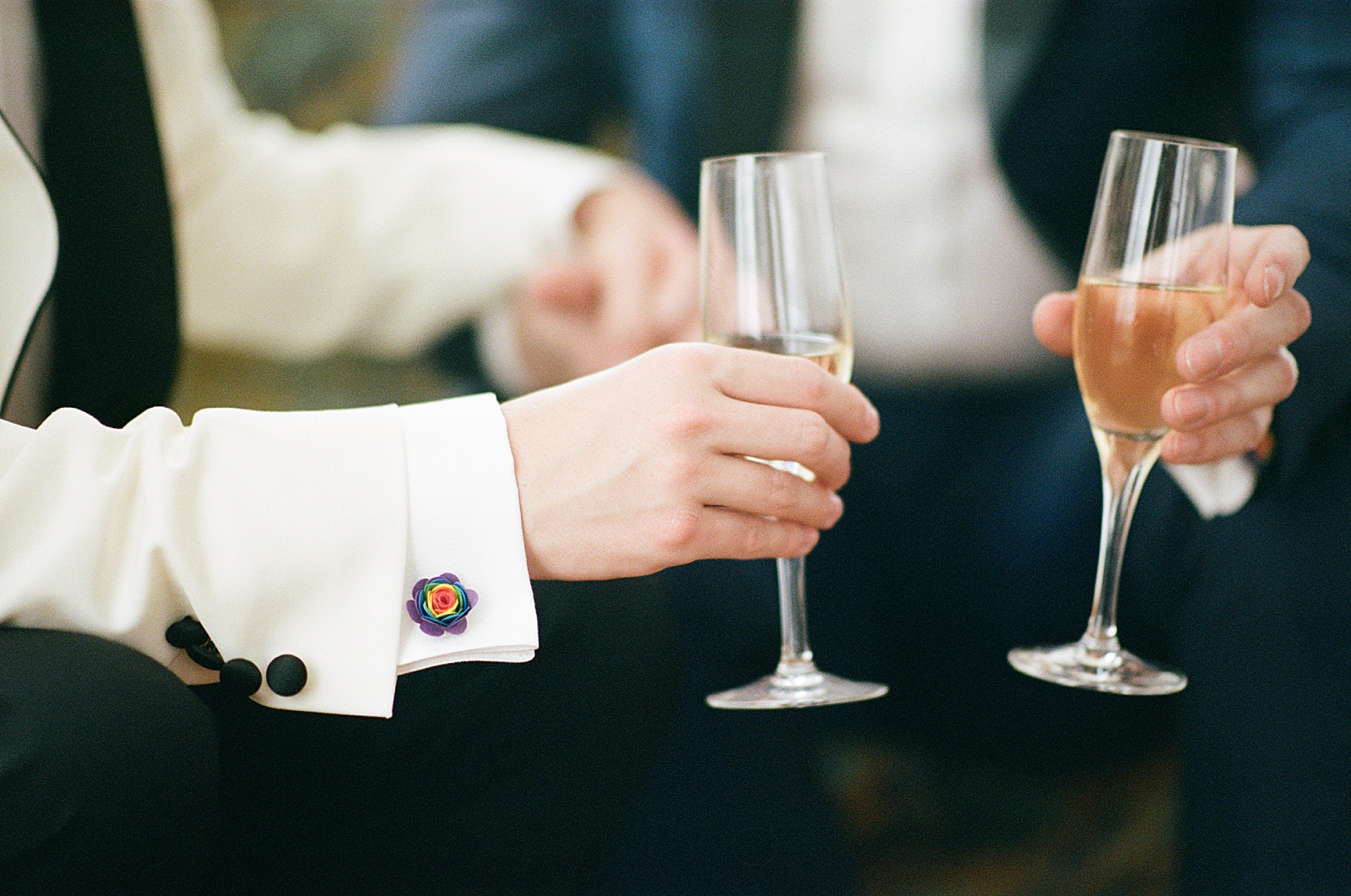 Two grooms hold hands and champagne flutes in wedding portraits on film.
