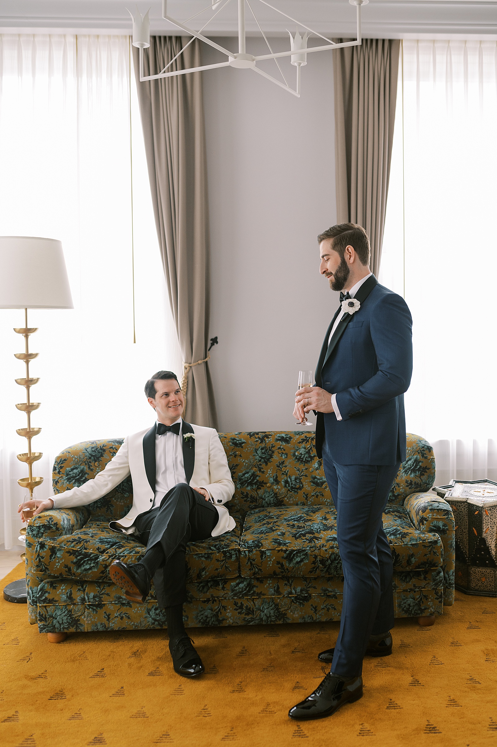 A groom stands beside another groom who is sitting on a couch while posing for wedding portraits.