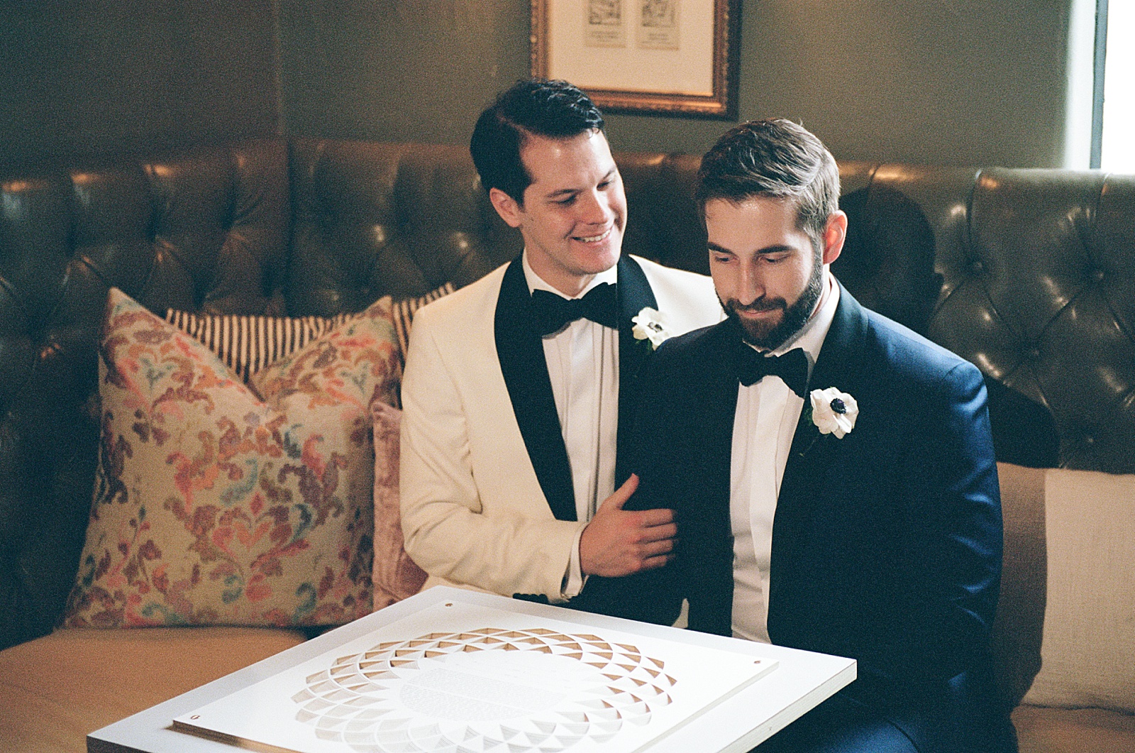 Two grooms sit next to each other on a couch looking at their ketubah.