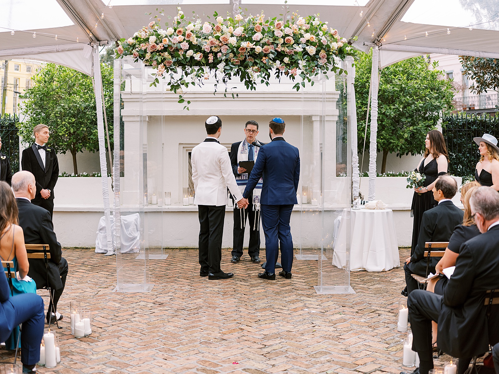 Two grooms hold hands under a floral chuppah at a wedding in a courtyard.
