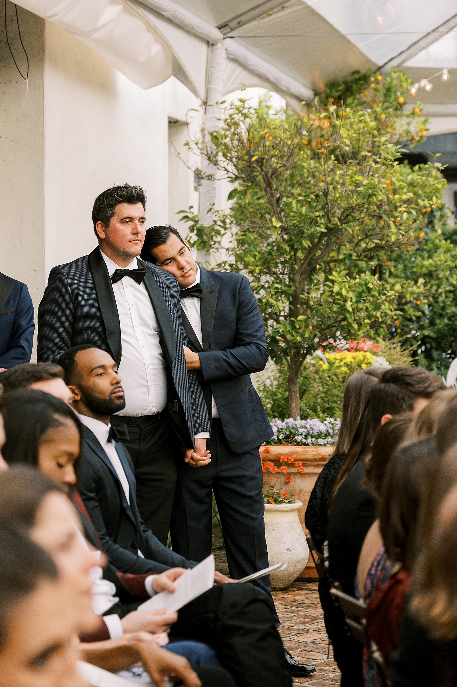 Two wedding guests lean on each other while they watch a wedding ceremony at a historic wedding venue.