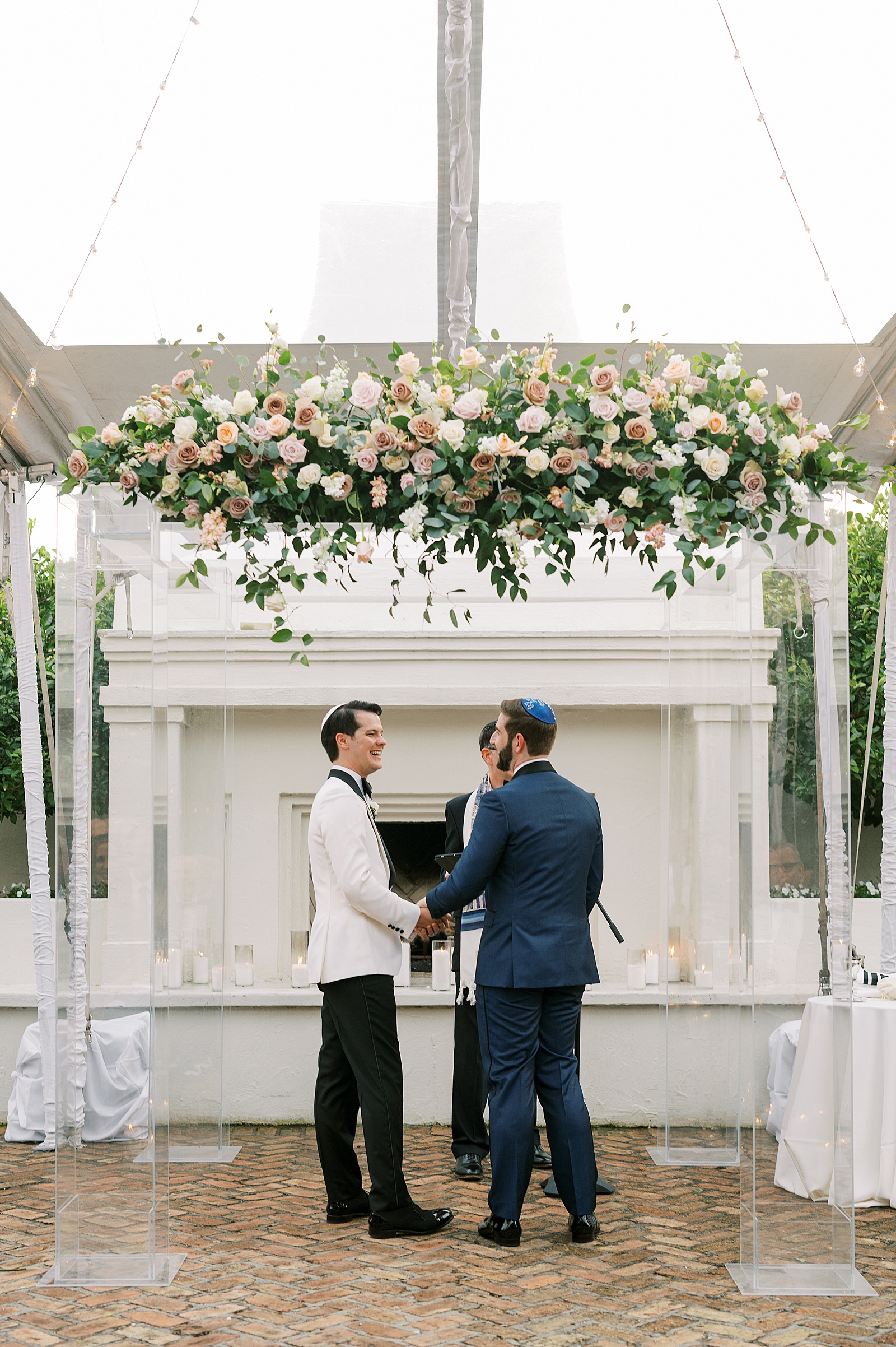 Two grooms turn to face each other under a floral chuppah in a courtyard wedding ceremony.