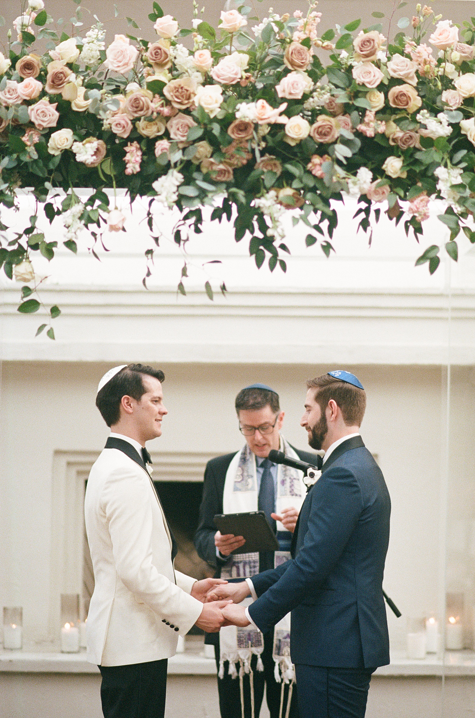 Two grooms hold hands under a floral chuppah during a Jewish wedding.