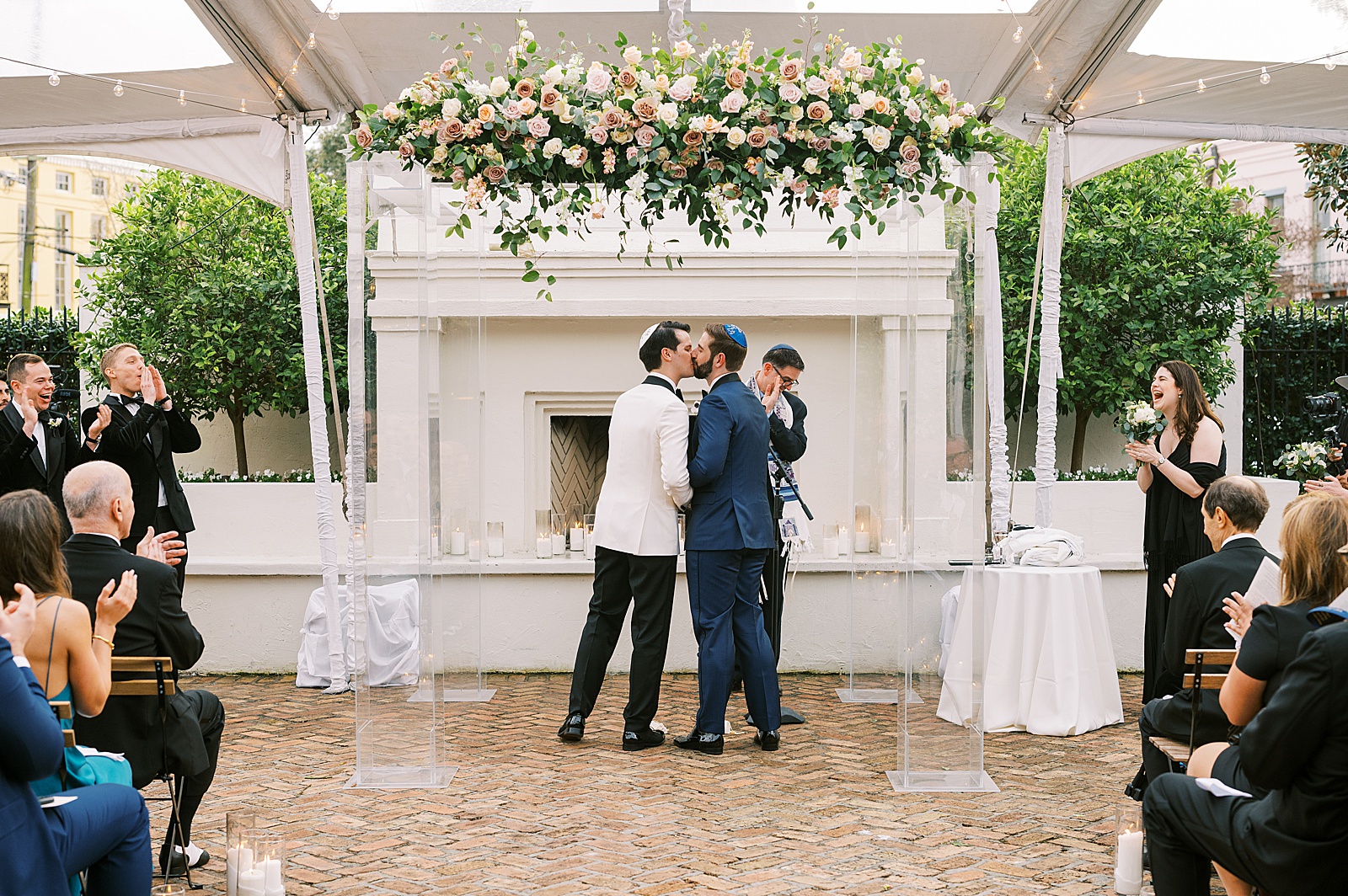 Two grooms kiss at the end of a Jewish wedding ceremony in a courtyard.