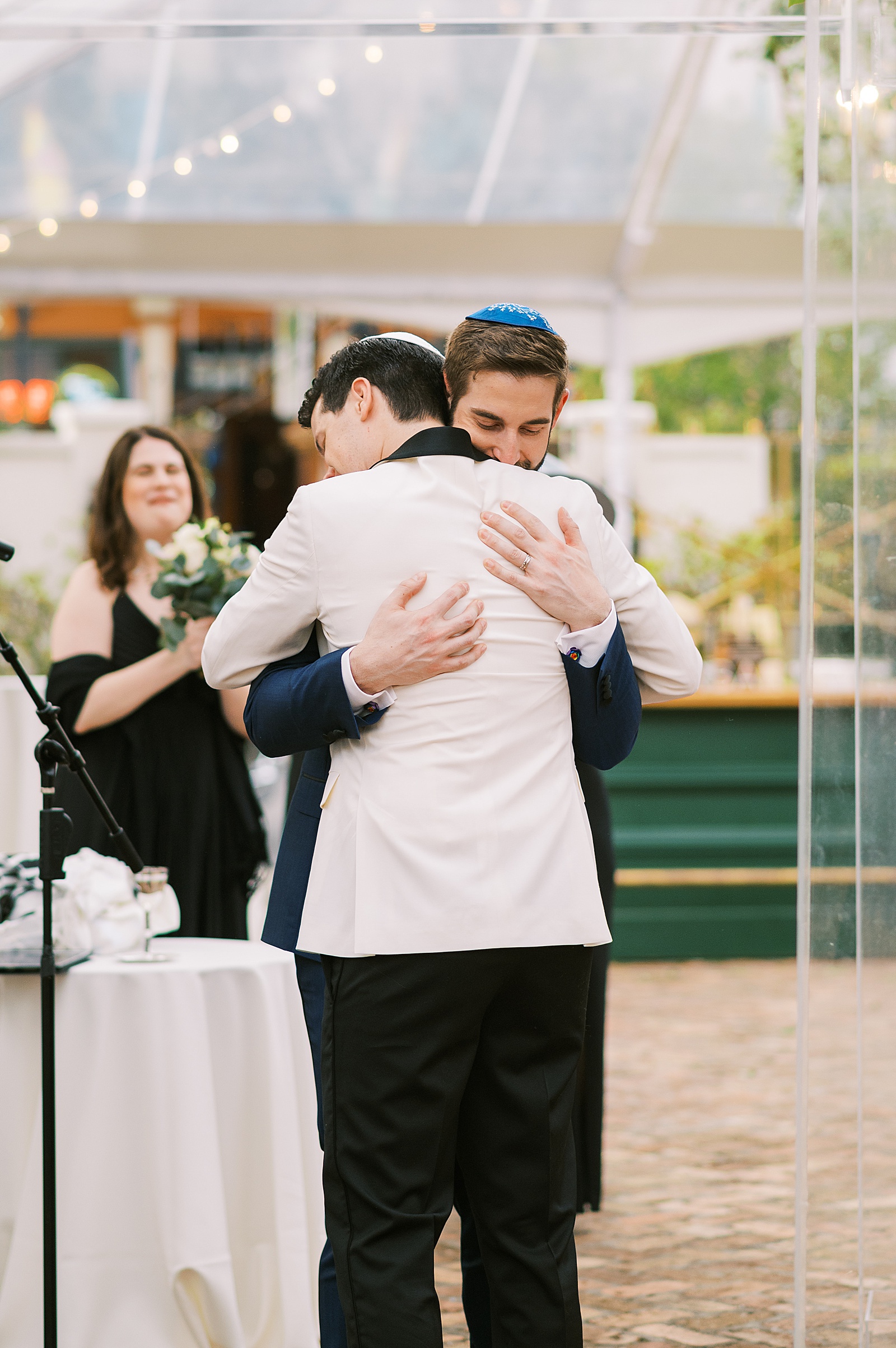 Two grooms embrace under a clear wedding tent in a candid wedding portrait on film photography.