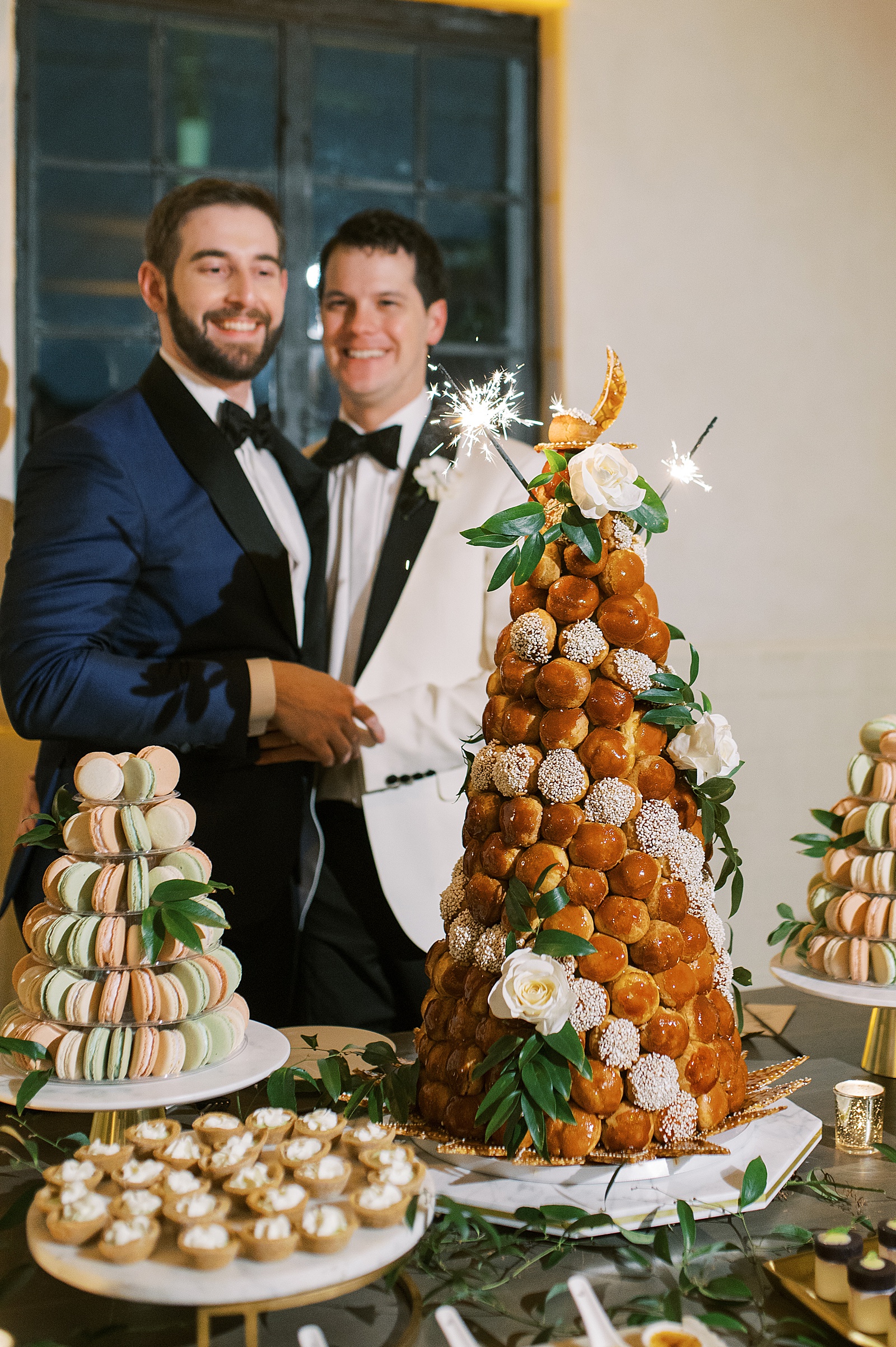 Two grooms stand behind a wedding croquembouche at their reception.