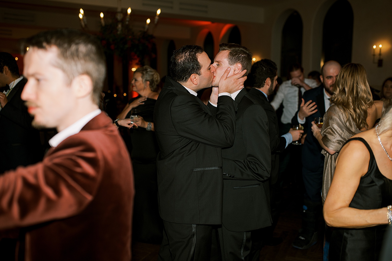 Two men kiss while people dance around them at a wedding reception.