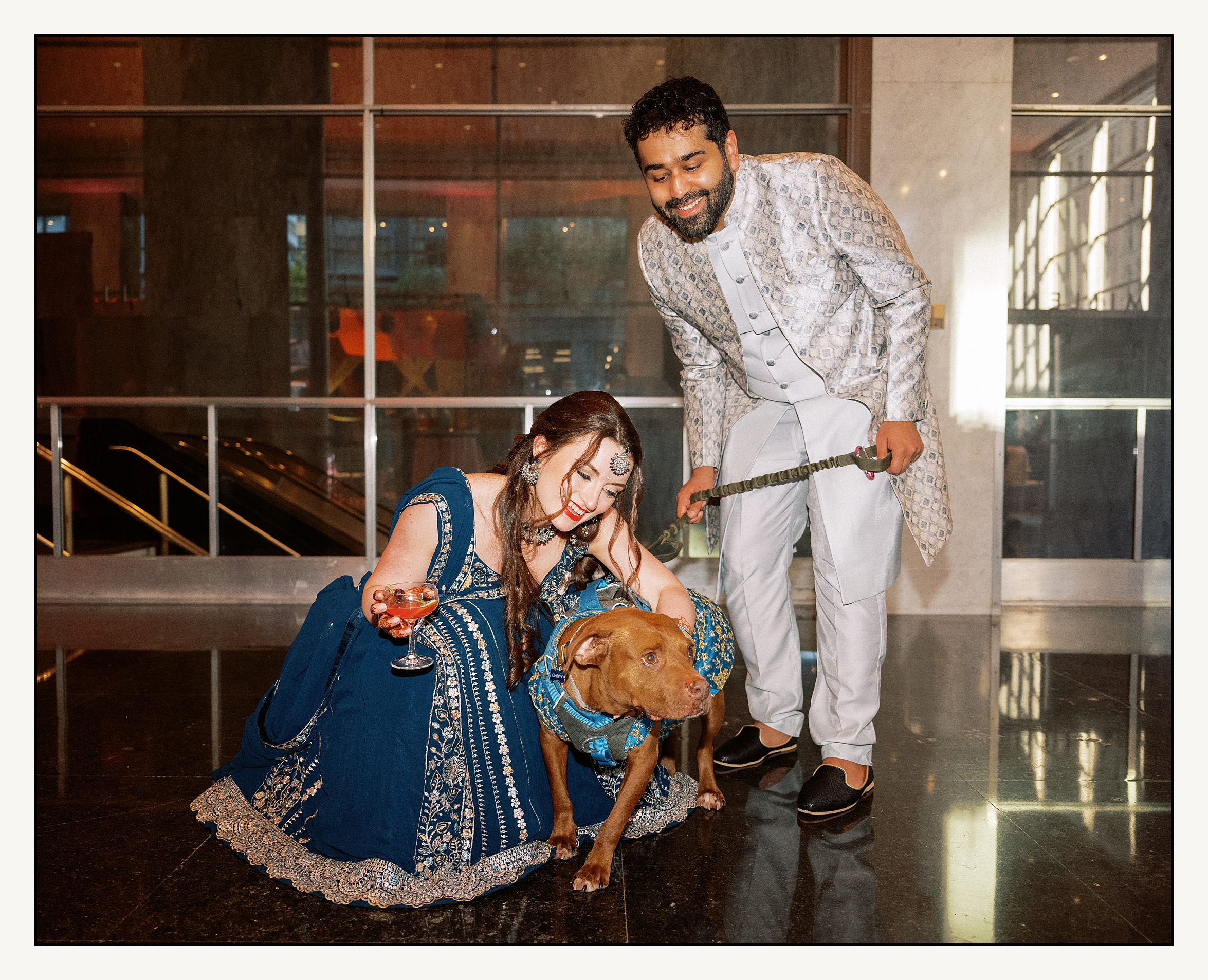 A bride and groom crouch down to hug their dog who is dressed in a dog wedding outfit.