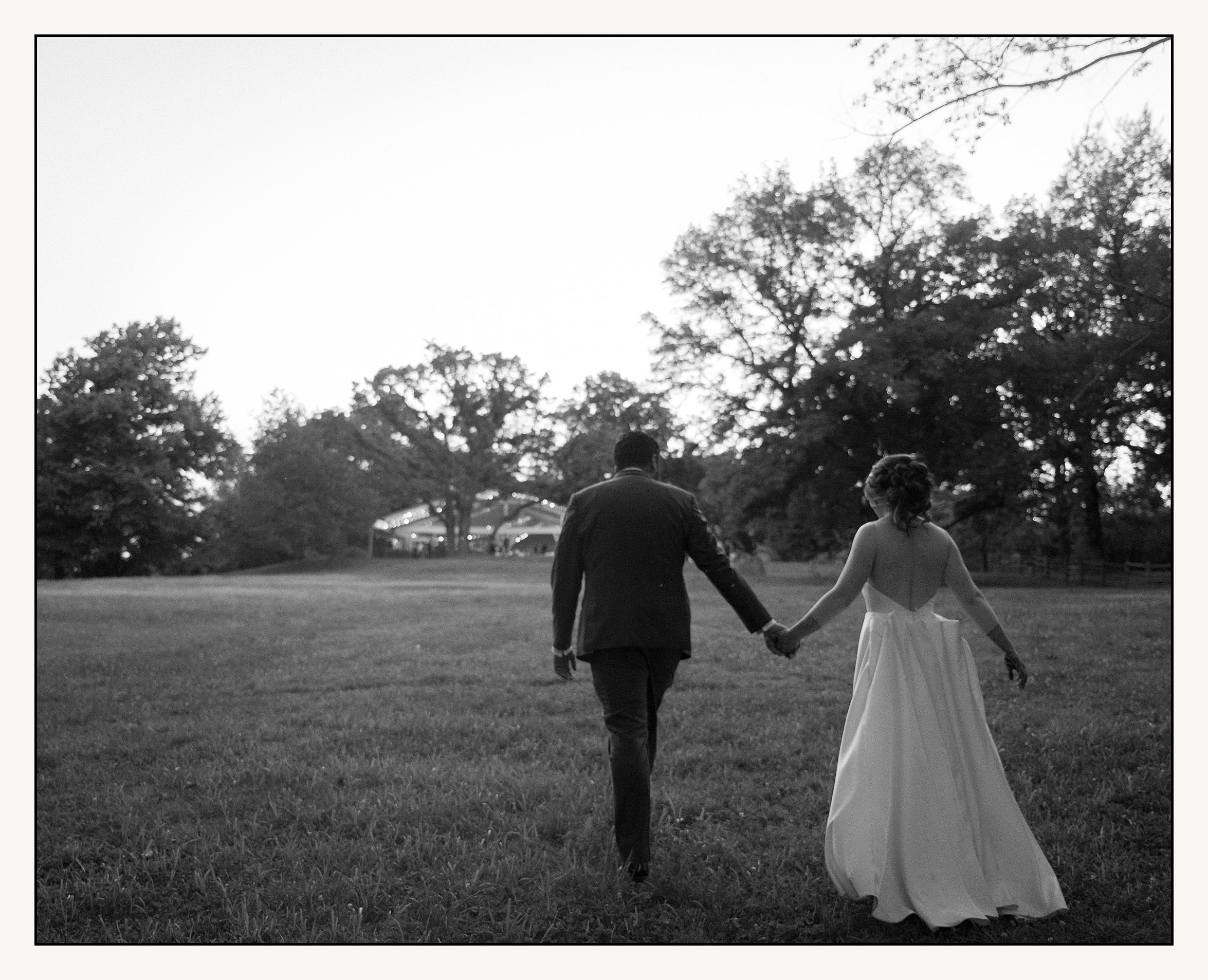 A bride and groom walk through a field towards a wedding tent at Bartram's garden.