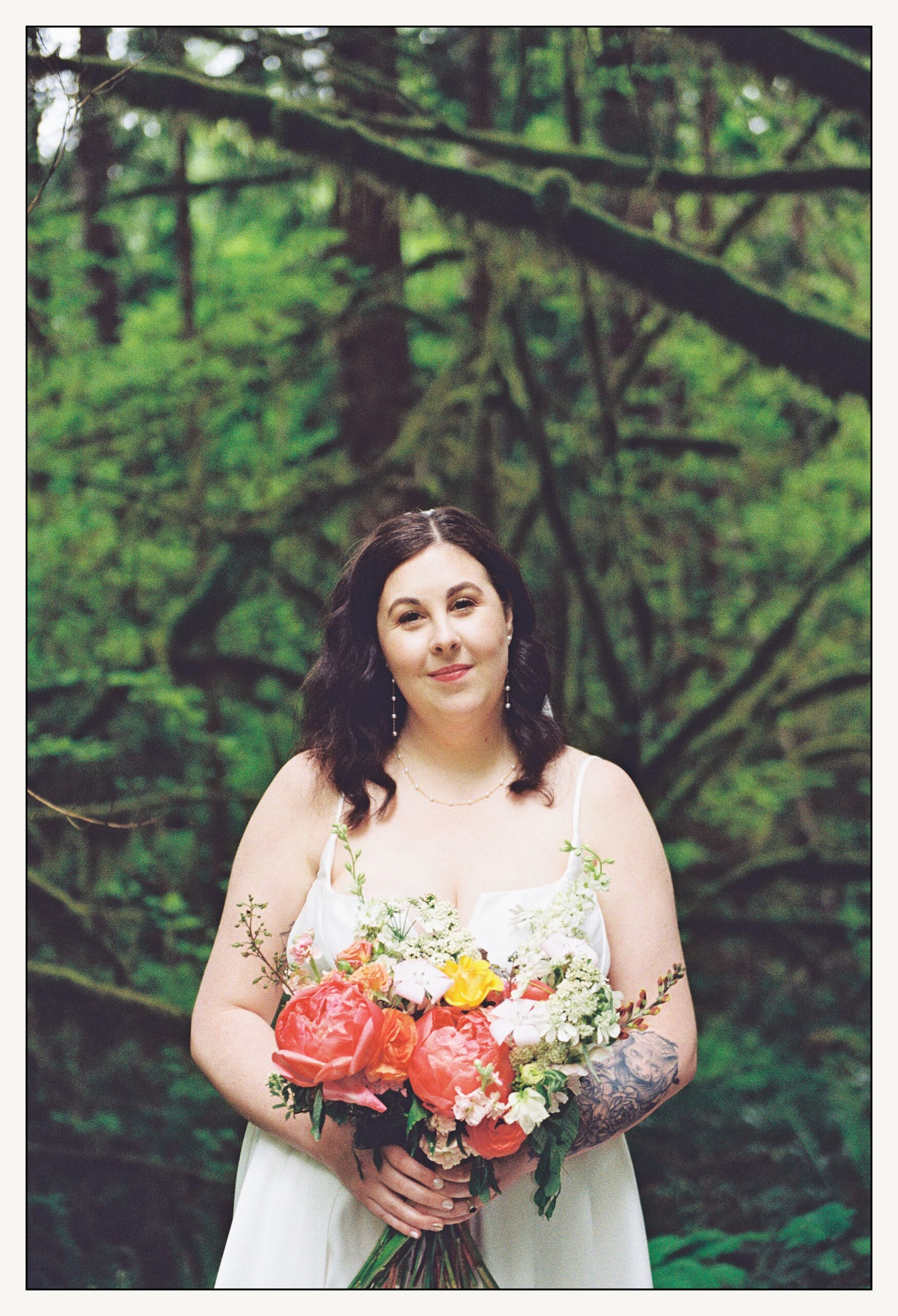 A bride stands in a Washington forest holding a bright pink bouquet for her PNW wedding.